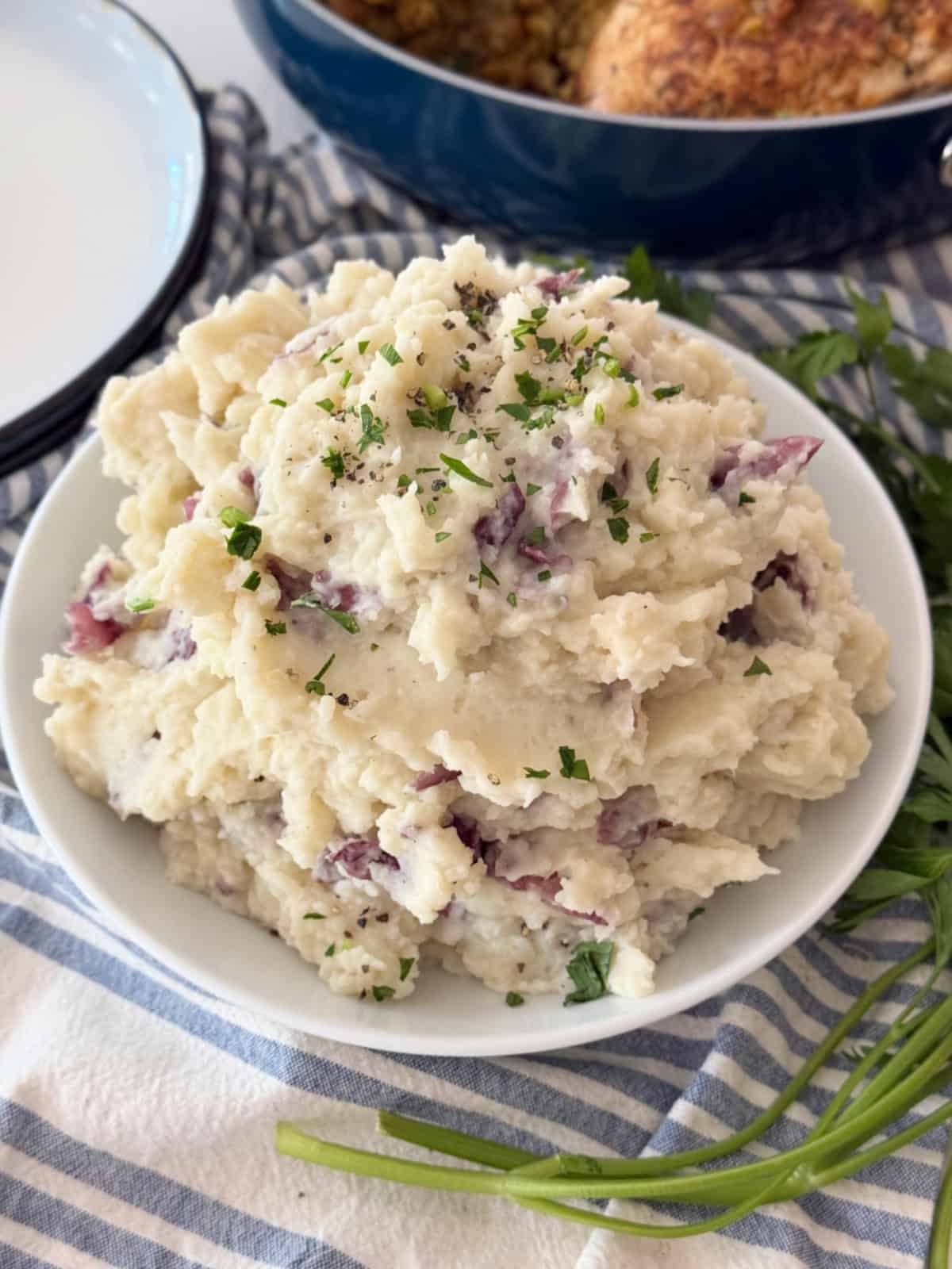 White bowl filled with mashed potatoes topped with parsley with chicken in blue pan in background.