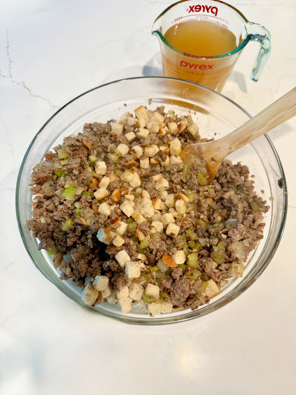 Glass bowl on a white marble counter filled with ground sausage and stuffing cubes.