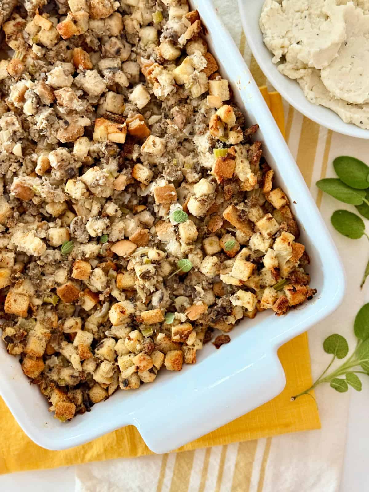 Sausage stuffing in a white baking dish on a yellow cloth next to a bowl of mashed potatoes.