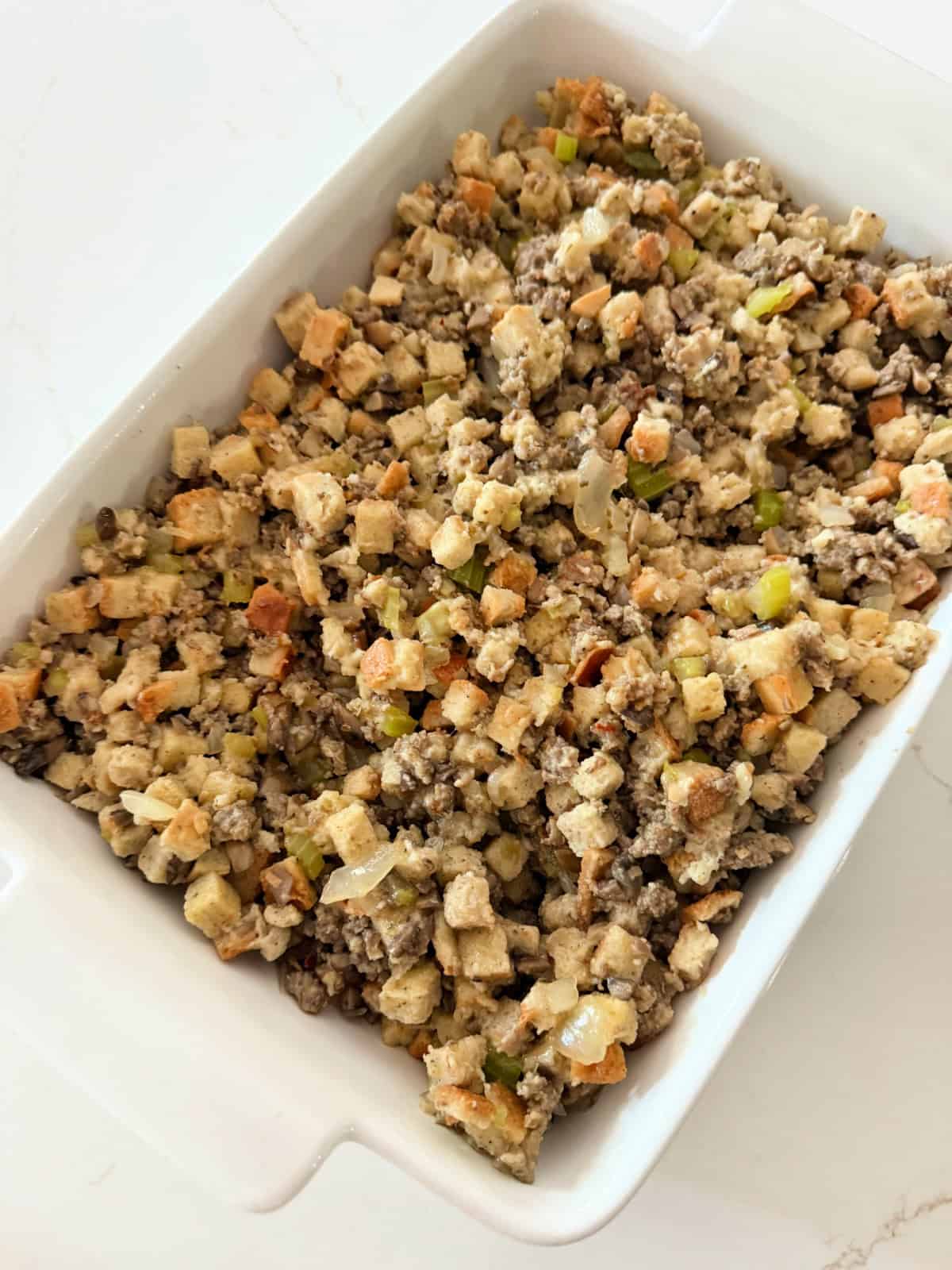 Uncooked sausage stuffing in a white baking dish on a white marble counter.