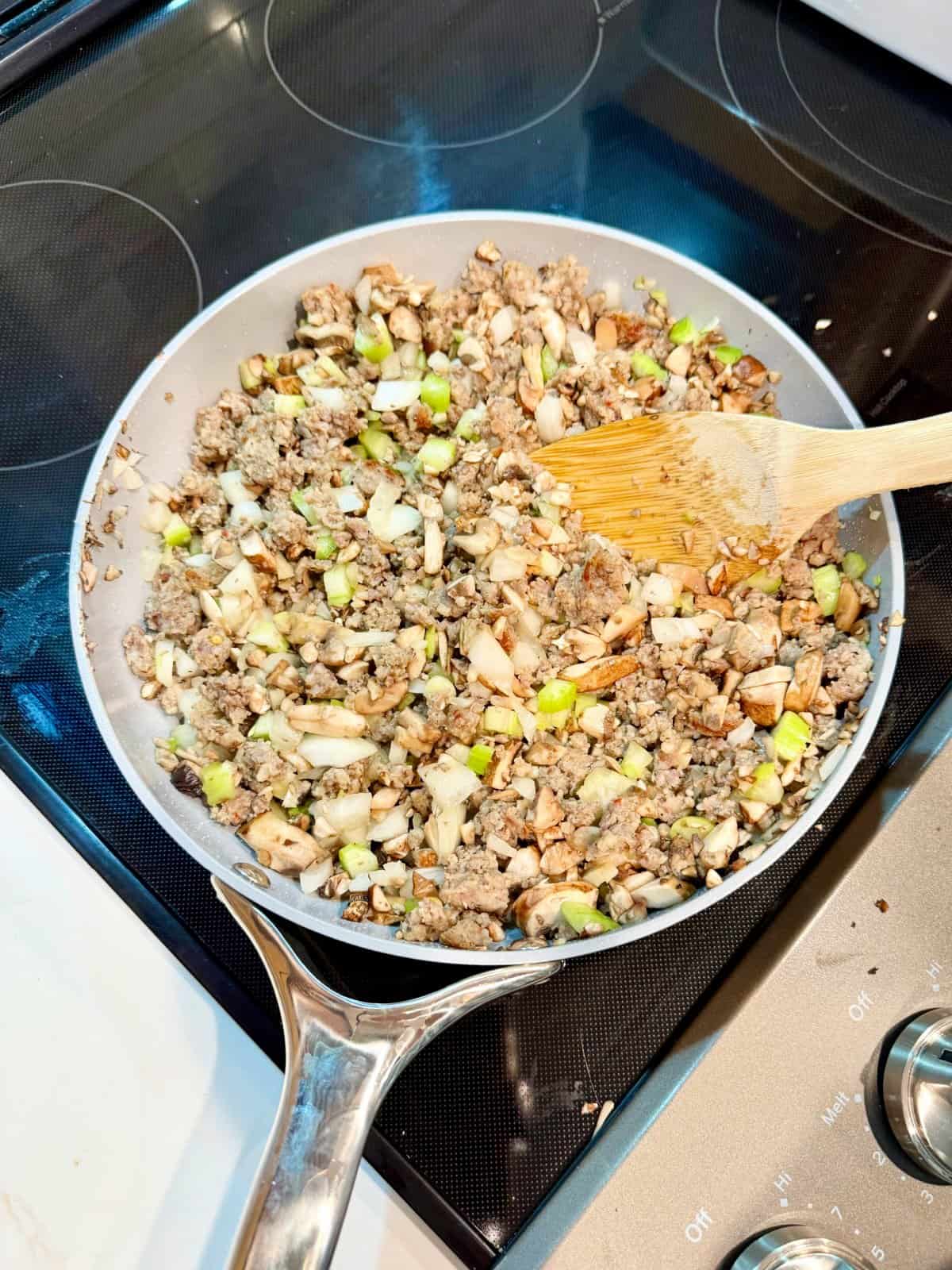 Frying pan on glass stove top filled with ground sausage, celery, onion, and chopped mushrooms.