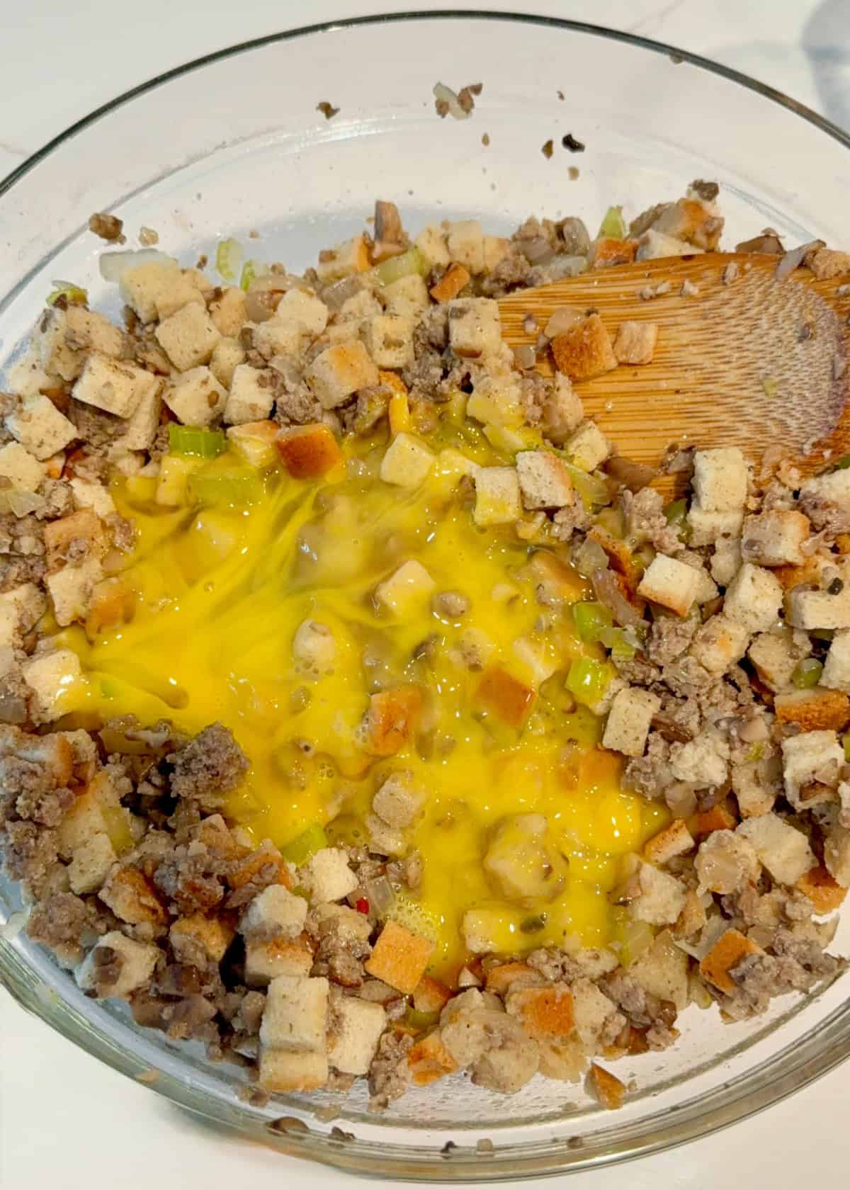 Glass bowl filled with cooked ground sausage, stuffing cubes, and eggs with a wooden spatula.