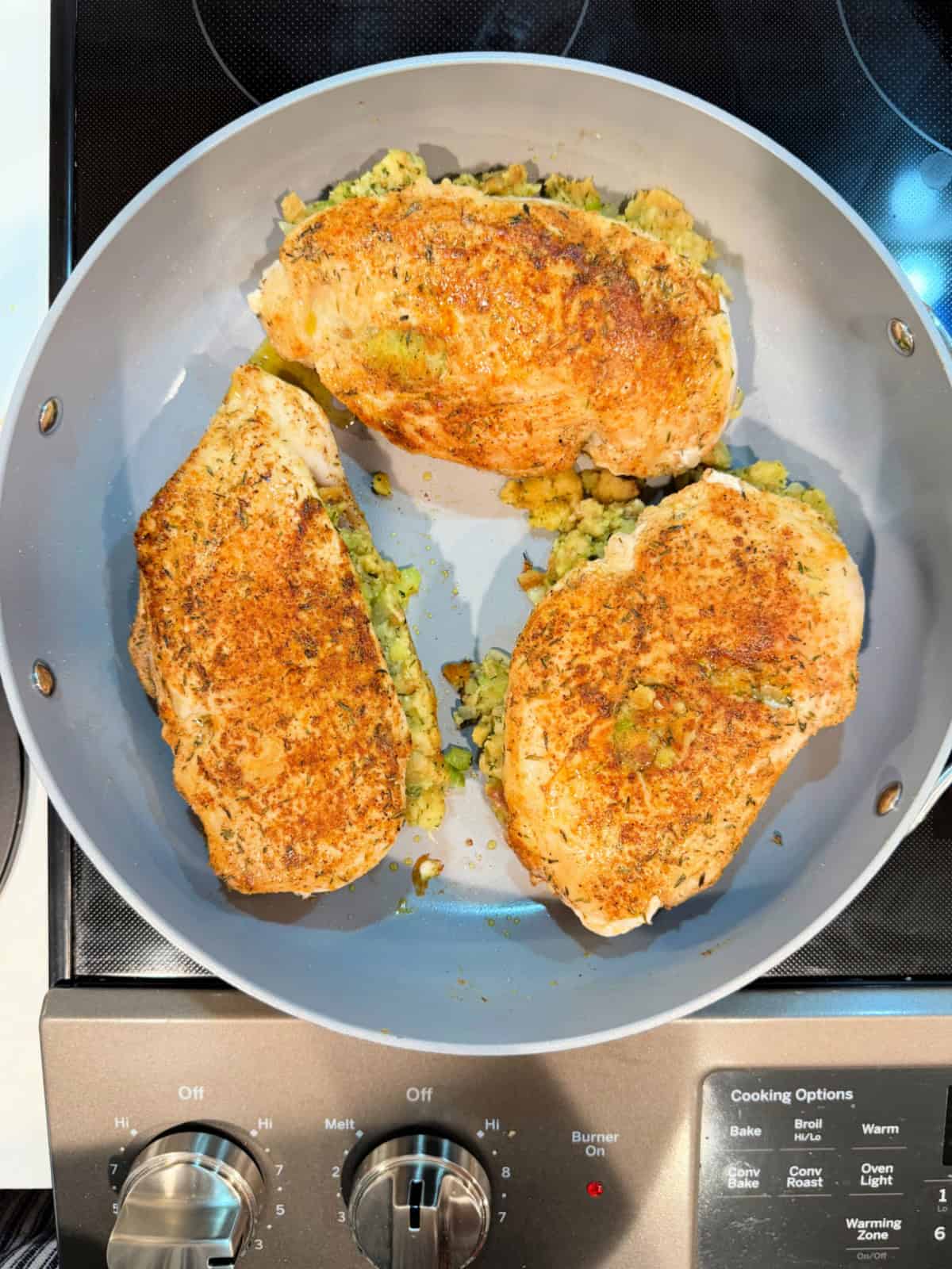 Gray frying pan with three browned chicken breast on the pan stuffed with stuffing on a stove top.