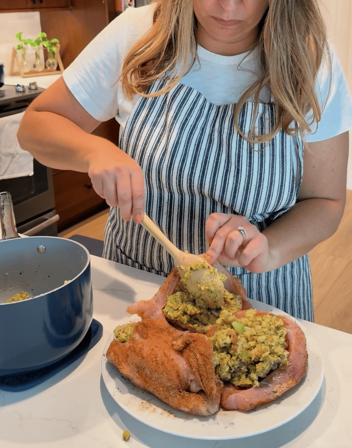 Female in apron holding open a split raw chicken breast holding a spoon and placing stuffing inside the chicken.