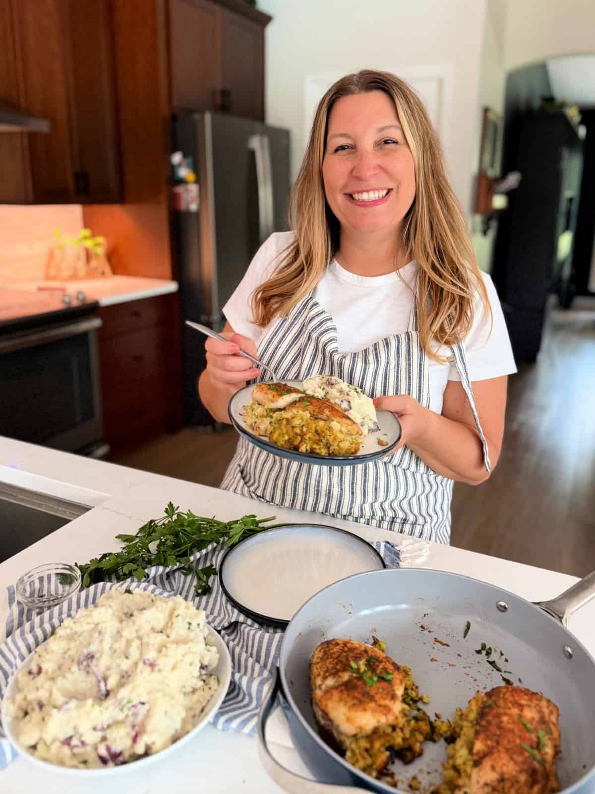Female with an apron on standing in a kitchen holding a plate of food with mashed potatoes in chicken.