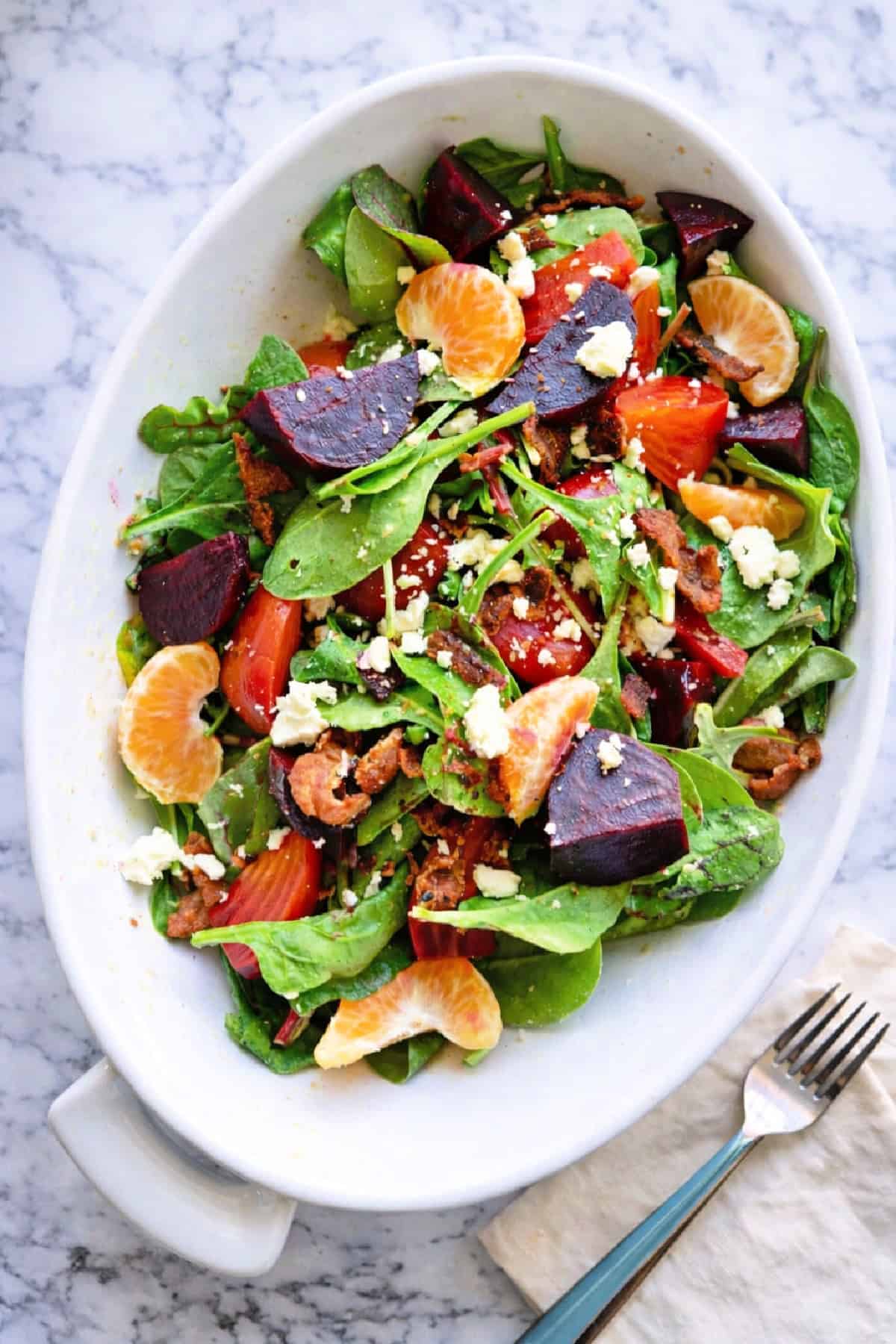 White oval serving dish filled with a citrus and beet salad on a marble tabletop with a cream napkin and forks.