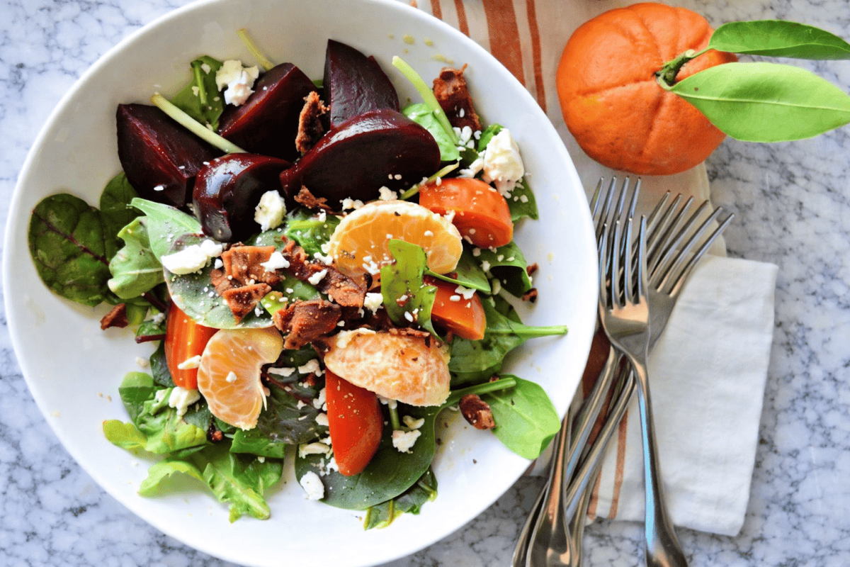 White bowl with leafy green salad with beets, oranges, bacon and cheese on a marble countertop with forks and an orange next to it.