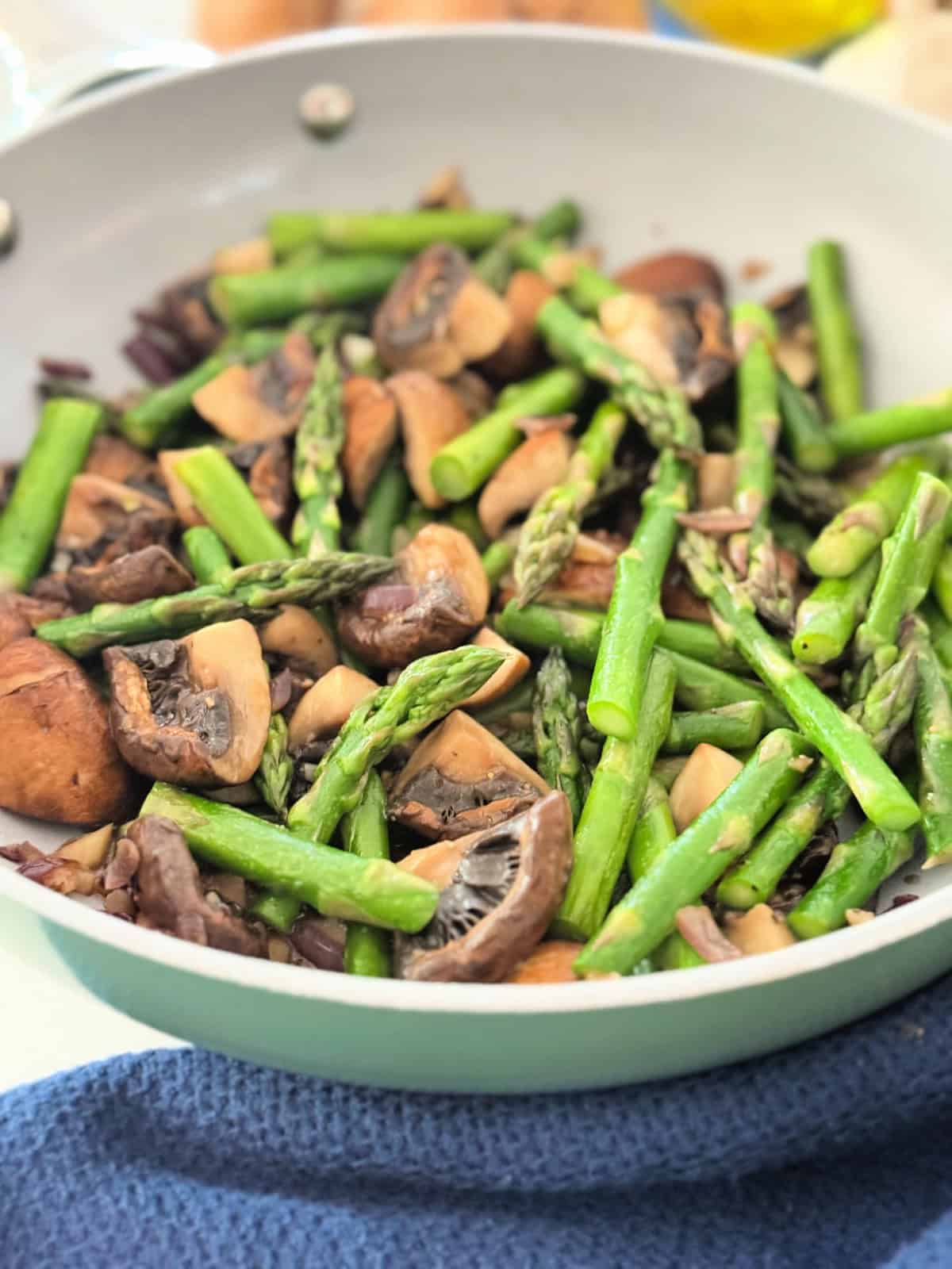 Pieces of asparagus and mushrooms in a green frying pan on a white countertop with blue cloth.